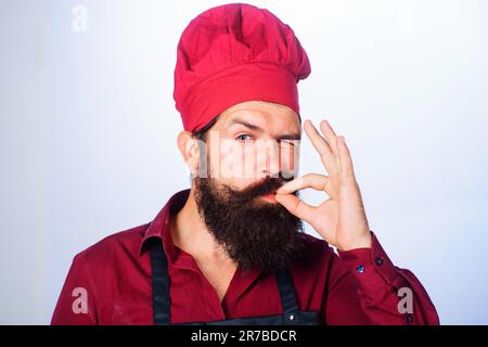 Restaurantwerbung. Professioneller Koch mit Schild für Delicious. Der zuzwinkernde Koch, der Koch oder der Bäcker waren ausgezeichnet. Männlicher Koch mit perfektem Schild Stockfoto