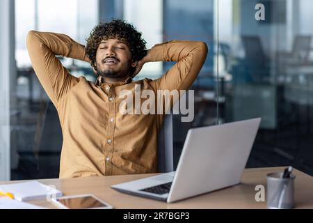 Aufgabe erfolgreich abgeschlossen und gut gemacht, Geschäftsmann tippt im Büro, hispanischer Mann mit Händen hinter dem Kopf, träumte und lächelte mit geschlossenen Augen. Stockfoto