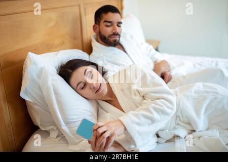 Arabischer Ehemann, der die Nachrichten seiner Frau auf dem Handy liest, liegt im Schlafzimmer Stockfoto