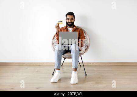 Mann mit Kreditkarte und Laptop, der an der weißen Wand sitzt Stockfoto