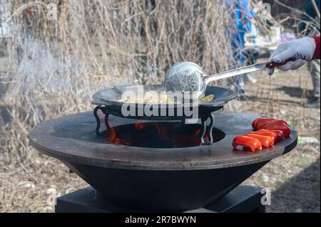 Gemüse wird auf einem runden Herd geröstet. Runder Grill in Form einer Schüssel mit Feuer im Inneren. Mann bereitet Herd für Barbecue zu. Das Brennholz brennt Stockfoto