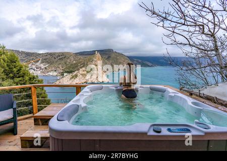 Nehmen Sie sich Zeit für sich. Badesachen im Freien mit Blick auf die Berge und das Meer. Eine Frau im schwarzen Badeanzug entspannt sich im Hotelpool und bewundert die Aussicht Stockfoto