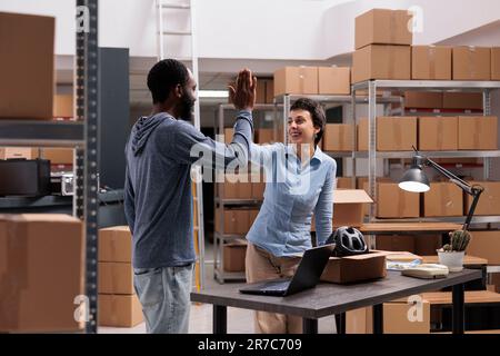 Glückliche Lagermitarbeiter haben High Five geschafft, nachdem sie Helmpakete in Pappkartons verpackt haben, Arbeiter, die im Warenlager arbeiten. Lächelnde Leute, die ihren Triumph im Lagerhaus feiern Stockfoto