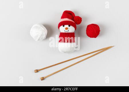 Gestrickter Schneemann mit rotem Hut und Pullover mit Fäden und Stricknadeln auf grauem Hintergrund. Babysachen und Accessoires. Geschenk für den Winter Stockfoto