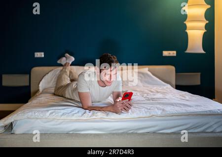 Mann liest Informationen auf Smartphone, liegt zu Hause im Bett im modernen Schlafzimmer am Abend. Stockfoto