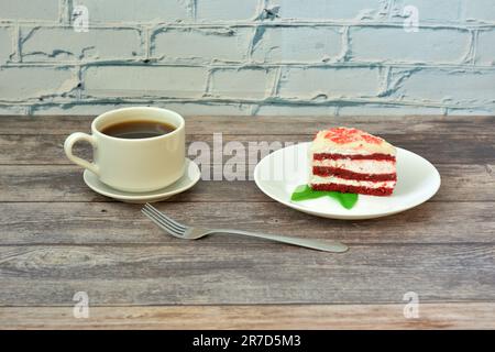 Eine Tasse heißen schwarzen Kaffee auf einer Untertasse und ein Teller mit einem Stück roten Samtkuchen auf einem hellen Holztisch. Nahaufnahme. Stockfoto