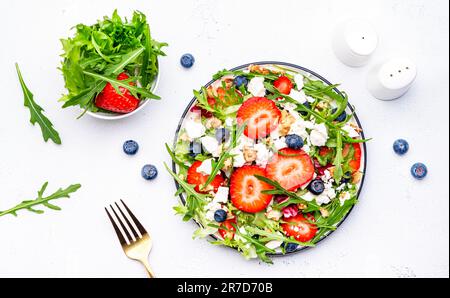 Erdbeersalat mit Rucola, Kopfsalat, Blaubeeren, Fetakäse und Walnüssen, weißer Tisch. Frisches, nützliches Gericht für gesunde Ernährung Stockfoto