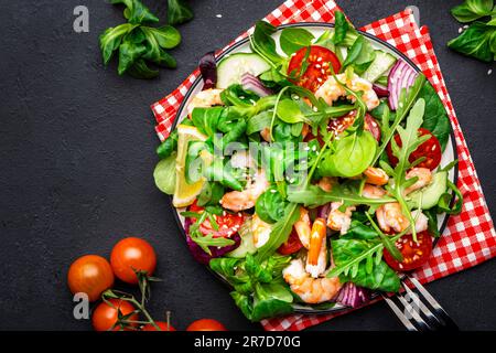 Leckerer Salat mit Avocado, Garnelen, roten Tomaten, Gurke, Rucola, Feldsalat, Zwiebeln und Sesamsamen auf schwarzem Tischhintergrund. Draufsicht, Kopie Spa Stockfoto