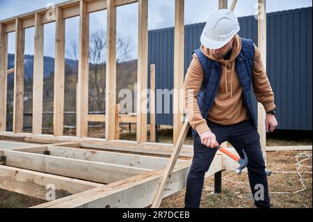 Mann Arbeiter Gebäude Holzrahmenhaus auf Pfahlfundament. Der Zimmermann hämmert mit Hammer den Nagel in die Holzdiele. Schreinerei-Konzept. Stockfoto