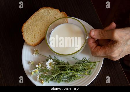 Die alte Hand hält eine Tasse Milch in der Nähe von Gänseblümchen und ein Stück Brot an einem Tisch Stockfoto