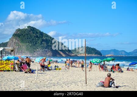 Rio de Janeiro, Brasilien - 25. Mai 2023: Strandbesucher genießen einen sonnigen Tag am Copacabana-Strand unter Sonnenschirmen. Das Meer und der üppige grüne Berg Stockfoto