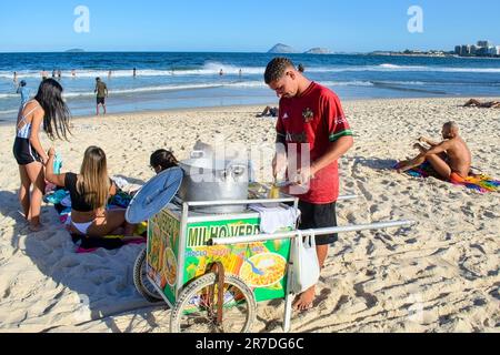 Rio de Janeiro, Brasilien - 25. Mai 2023: Ein Mann steht hinter einem Karren am Strand von Copacabana und macht Popcorn in einem offenen Topf. Die Leute entspannen sich im sandigen Bauch Stockfoto