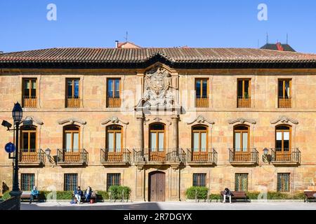 Oviedo, Spanien - 12. Februar 2023: Leute sitzen auf Bänken vor einem geschossigen Gebäude mit Balkonen und Glasfenstern. Die Struktur zeigt ein Stockfoto