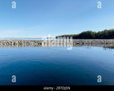 Das ruhige Wasser eines Sees trifft auf eine felsige Barriere unter einem klaren blauen Himmel. Stockfoto