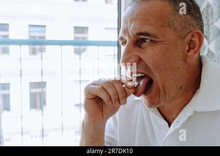 Ein Mann mittleren Alters, braunäugig, in weißem Polo, sitzt auf einem Stuhl am Fenster, nimmt Pillen. Stockfoto