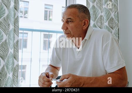 Ein Mann mittleren Alters, braunäugig, in weißem Polo, sitzt auf einem Stuhl am Fenster, nimmt Pillen. Stockfoto