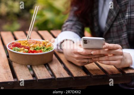 Nahaufnahme einer Frau, die ihr Handy benutzt, eine Schüssel mit Gemüse, Obst und Essstäbchen auf dem Tisch. Park mit viel Grün Stockfoto