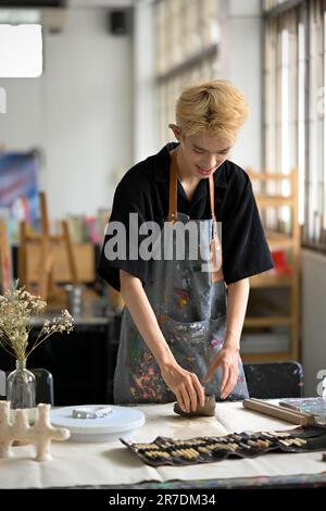 Ein Porträt eines jungen, asiatischen, schwulen, männlichen Künstlers oder Töpfers in einer Schürze, der rohen Ton knete und Tonornamente an seinem Schreibtisch in seinem Töpferstudio herstellte. Stockfoto