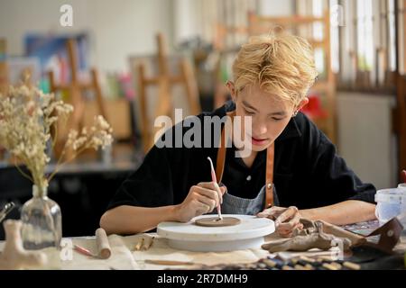 Ein talentierter und attraktiver junger asiatischer schwuler Töpferkünstler mit einem Bildhauer-Werkzeug konzentriert sich auf das Erstellen von Mustern auf rohem Ton in seinem Studio. Kunsthandwerker, Pot Stockfoto
