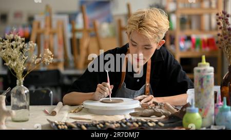 Ein kreativer und talentierter junger asiatischer schwuler Töpferkünstler mit einem Bildhauer-Werkzeug konzentriert sich in seinem Studio auf das Erstellen von Mustern auf rohem Ton. Stockfoto