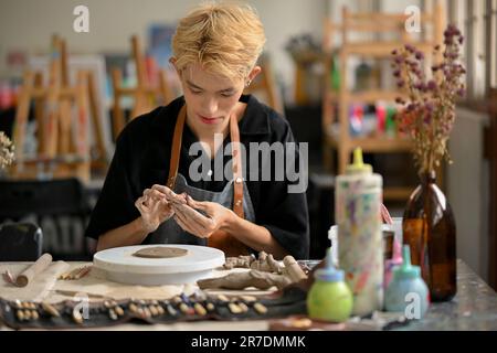 Ein fokussierter und talentierter junger asiatischer, schwuler Künstler, der rohen Lehm formt, um ein handgemachtes Schmuckstück zu machen, und in seinem Studio arbeitet. Stockfoto