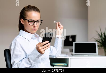Seriöse junge Geschäftsfrau mit Brille, die am Schreibtisch in ihrem eigenen Büro sitzt und ihr Smartphone benutzt. Stockfoto