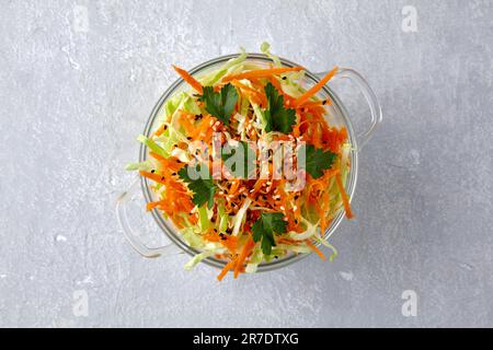 Heller Sommersalat aus Weißkohl und Karotten mit Sesam und schwarzen Kreuzkümmelsamen in einer Glasschüssel auf steingrauem Hintergrund. Draufsicht mit Kopierbereich Stockfoto