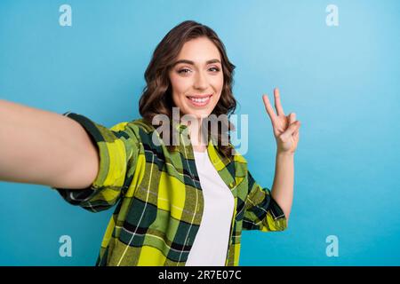 Porträt von positiv fröhlich niedlichem Mädchen wellige Frisur Kleidung karierte Jacke mit Selfie mit V-Zeichen isoliert auf blauem Hintergrund Stockfoto