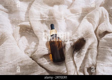 Braune Serumflasche und trockene Distelblüte auf beigefarbenem Baumwollgewebe-Hintergrund bei Sonnenlicht. Draufsicht. Stockfoto