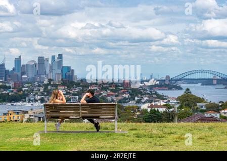 Ein junges Paar, das die Aussicht ignoriert, sitzt auf einer Parkbank mit Blick auf den Hafen von Sydney, die Brücke und das Opernhaus von Sydney Stockfoto