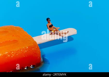 Eine Miniaturfrau in einem schwarzen Bikini trägt Sonnencreme auf ihre Beine auf, während sie auf dem Stab eines schmelzenden orangefarbenen Eis am Stiel auf einem blauen Backgr sitzt Stockfoto