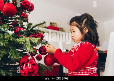 Bezauberndes 3-jähriges Kleinkind, das den Weihnachtsbaum dekoriert und einen roten Pullover trägt Stockfoto