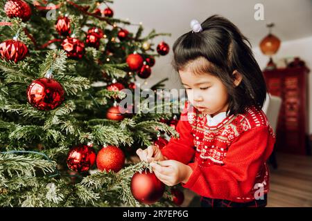 Bezauberndes 3-jähriges Kleinkind, das den Weihnachtsbaum dekoriert und einen roten Pullover trägt Stockfoto