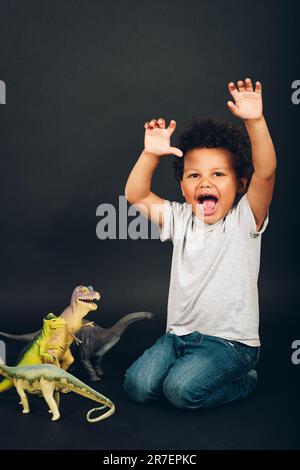 Studioaufnahme eines niedlichen afrikanischen 2-3-jährigen Kleinkindes, der mit Dinosaurierfiguren spielt, dunkler Hintergrund Stockfoto