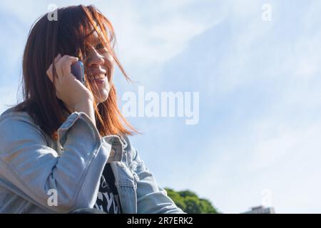 Junge Latina-Frau kolumbianischer Herkunft, die draußen sitzt und mit ihrer Familie telefoniert, mit dem Himmel im Hintergrund und Kopierraum Stockfoto