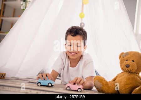 Foto eines niedlichen Vorschuljungen im heimwerkerlager, der Spielzeugautos spielt Stockfoto