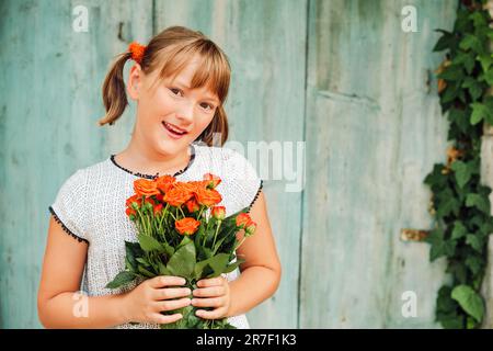 Außenporträt eines kleinen yong-Mädchens mit 9 Jahren, das ein weißes Kleid trägt und einen frischen Strauß wunderschöner orangefarbener Rosen hält Stockfoto