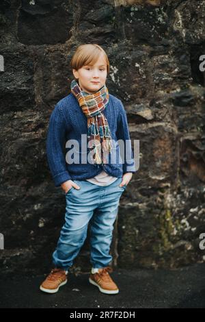 Außenporträt eines hübschen kleinen Jungen mit Jeans, blauer Strickjacke und braunen Schuhen, stilvolles Kind, das gegen dunkle Wände posiert. Mode für SM Stockfoto