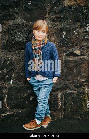 Außenporträt eines hübschen kleinen Jungen mit Jeans, blauer Strickjacke und braunen Schuhen, stilvolles Kind, das gegen dunkle Wände posiert. Mode für SM Stockfoto