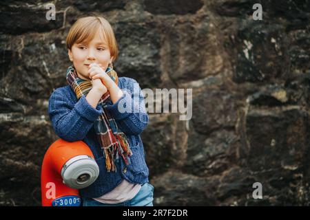 Außenporträt eines gutaussehenden kleinen Jungen mit Jeans, blauer Strickjacke, stilvollem Kind, das gegen dunkle Wände posiert. Mode für Kleinkinder Stockfoto