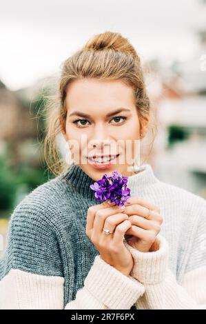 Frühlingsporträt einer hübschen jungen Frau, die einen warmen grauen Pullover trägt und einen kleinen Strauß Woolly Blue Violet hält (Viola sororia) Stockfoto