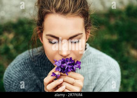 Frühlingsporträt einer hübschen jungen Frau, die einen warmen grauen Pullover trägt und einen kleinen Strauß Woolly Blue Violet hält (Viola sororia) Stockfoto