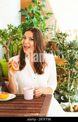 Junge, glückliche Frau, die auf dem Balkon frühstückt, Grapefruit oder rote Orange isst, eine Tasse Tee auf dem Tisch Stockfoto