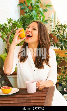 Junge, glückliche Frau, die auf dem Balkon frühstückt, Grapefruit oder rote Orange isst, eine Tasse Tee auf dem Tisch Stockfoto