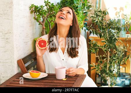 Junge, glückliche Frau, die auf dem Balkon frühstückt, Grapefruit oder rote Orange isst, eine Tasse Tee auf dem Tisch Stockfoto