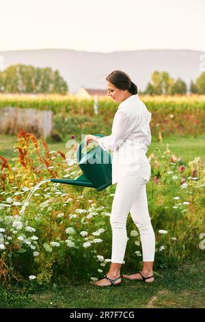 Außenporträt einer glücklichen Frau, die Pflanzen in einem wunderschönen Garten bewässert, weiße Kleidung und Hut trägt Stockfoto
