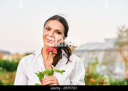 Nahaufnahme des Porträts einer schönen, reifen Frau, die einen schönen Tag im Garten genießt, mit rosa Zinnia-Blüten Stockfoto