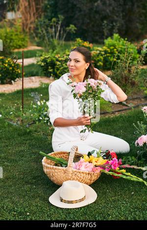 Gesunde, glückliche Frau, die im Blumengarten arbeitet, auf Gras sitzt, Dahlienblüten schneidet, Naturleben, Hobby und Freizeit Stockfoto