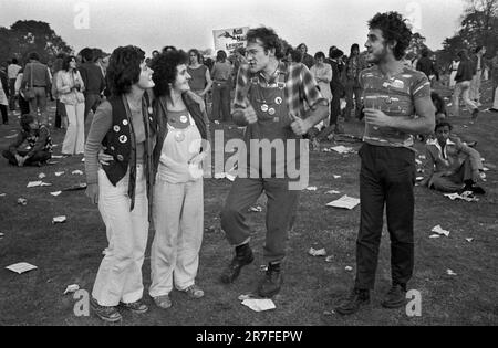 Fashion UK der 1970er Jahre, eine Gruppe junger Erwachsener, die trendige Latzhosen tragen, die mit Pin-Button-Abzeichen bedeckt sind, plaudern und Musik hören im Brockwell Park, im Rock Against Racism March and Concert. Sie marschierten vom Hyde Park zum Brockwell Park bei Brixton. Brockwell, South London, England 1978. HOMER SYKES Stockfoto