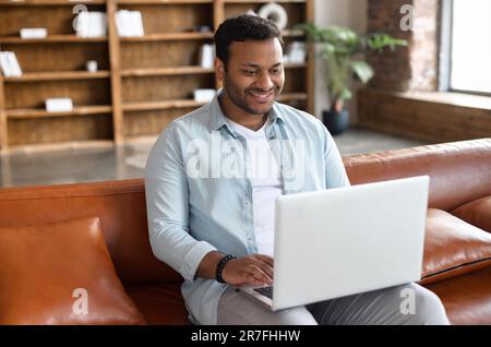 Ein fröhlicher, gut aussehender indianer in Freizeitkleidung, der auf dem Ledersofa mit Bücherregalen im Hintergrund sitzt, dunkelhaarige hispanische Männer im Internet surft, verbringt seine Freizeit in sozialen Netzwerken Stockfoto